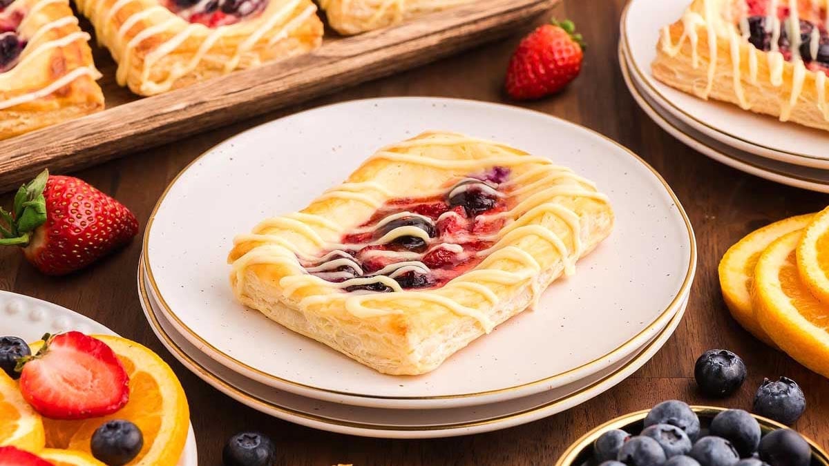 A rectangular fruit danish with icing is on a white plate, surrounded by strawberries, blueberries, and orange slices on a wooden table—a delicious testament to classic Danish recipes.