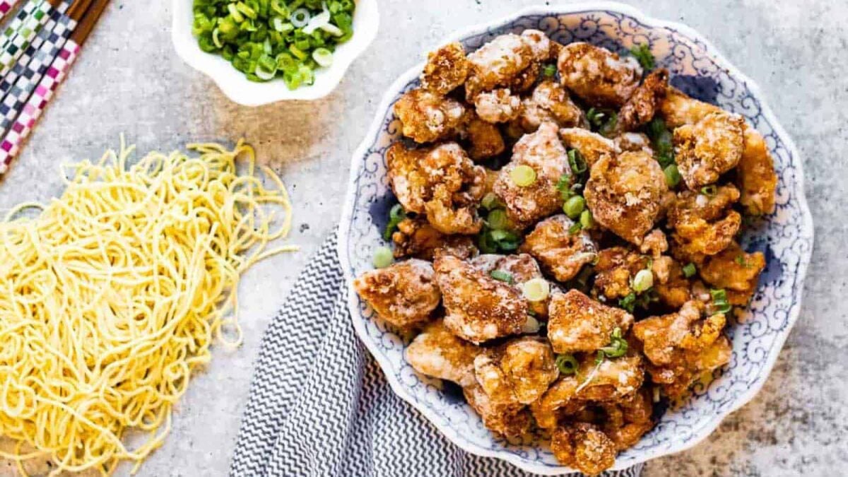 A bowl of crispy fried chicken pieces, an example of interesting chicken dishes, garnished with chopped green onions, sits beside a pile of dry noodles and a small bowl of sliced green onions.