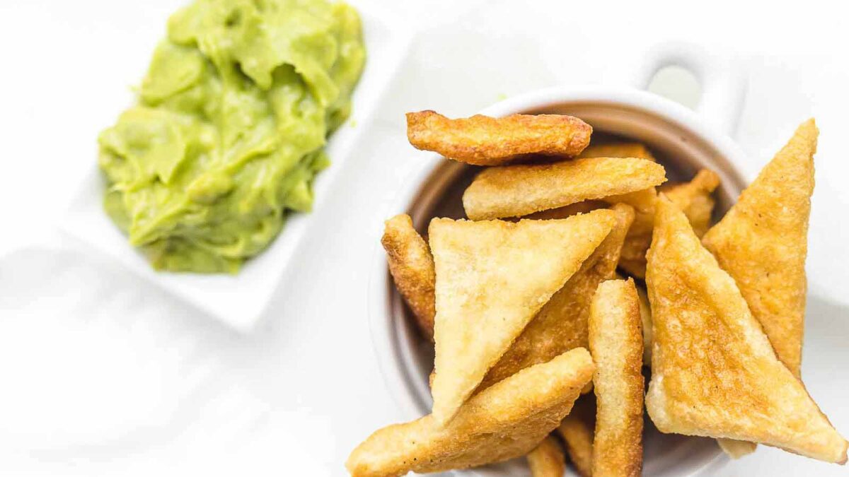 A white cup filled with crispy, golden crunch triangular chips sits next to a small dish of creamy guacamole on a white background.