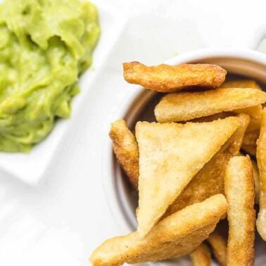 A white cup filled with crispy, golden crunch triangular chips sits next to a small dish of creamy guacamole on a white background.