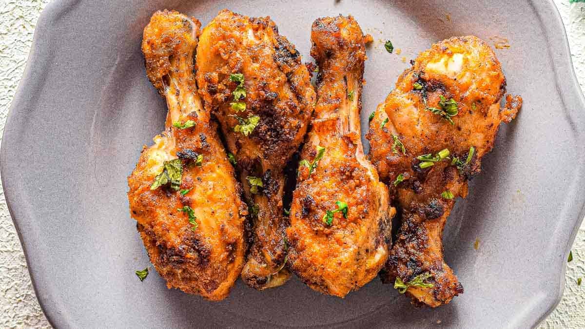 Three seasoned and baked chicken drumsticks with a golden crunch are garnished with chopped herbs and arranged on a gray plate.