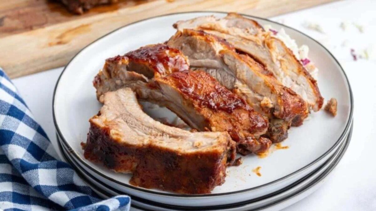 Four pieces of juicy, sauced pork ribs are arranged on a white plate, with a checkered napkin and wooden cutting board in the background.