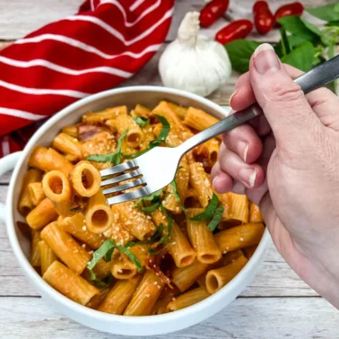 A hand holding a fork above a bowl of rigatoni pasta with tomato sauce and basil, styled to capture restaurant taste at home, with garlic, basil leaves, and tomatoes in the backgroundโperfect for trying copycat recipes.