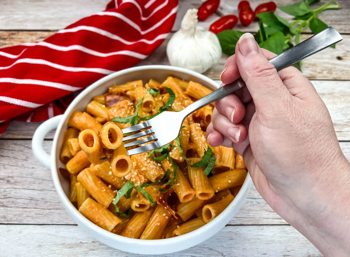 A hand holding a fork above a bowl of rigatoni pasta with tomato sauce and basil, styled to capture restaurant taste at home, with garlic, basil leaves, and tomatoes in the background—perfect for trying copycat recipes.