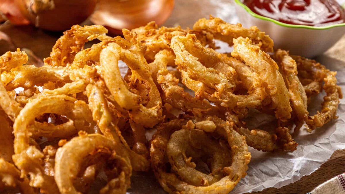 A pile of golden crunch fried onion rings on parchment paper with a bowl of ketchup in the background—the perfect addition to your favorite crispy recipes.