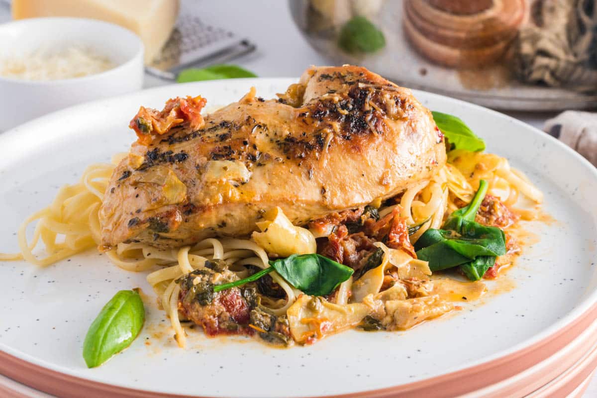 A cooked chicken breast served over pasta with a creamy sauce, sun-dried tomatoes, spinach, and fresh basil on a white plate—perfect for quick dinners and easy recipes.