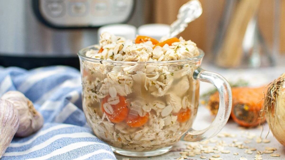 A clear glass mug filled with oatmeal, shredded chicken, sliced carrots, and herbs sits on a striped cloth near garlic, an onion, and a pressure cooker—perfect for easy Instant Pot meals.