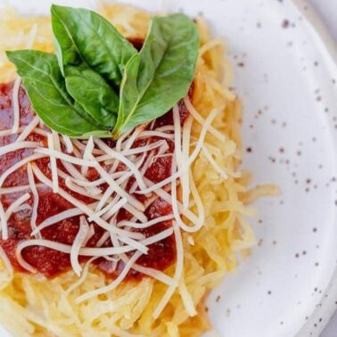 A plate of spaghetti squash topped with marinara sauce, shredded cheese, and fresh basil leaves, served on a speckled white plate—perfect for fans of light recipes.