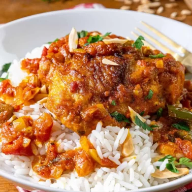 A plate of white rice topped with chicken in a flavorful curry tomato-based sauce, garnished with chopped herbs and sliced almonds, with a gold fork on the side.