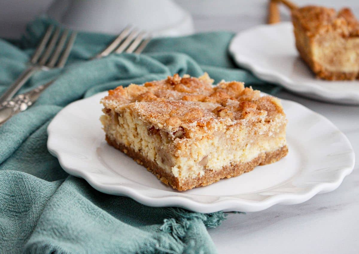 A square slice of crumbly Snickerdoodle dessert cheesecake bar on a white plate, with a hint of cinnamon-sugar, sits next to a teal napkin and forks.