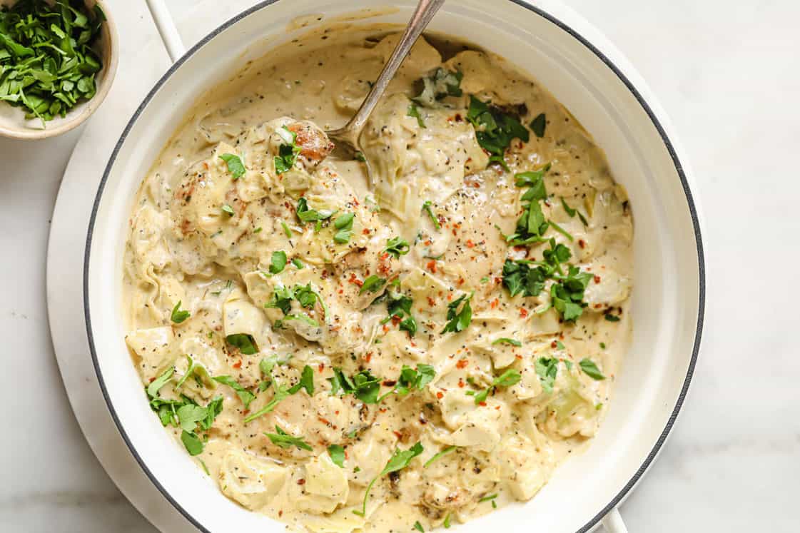 A bowl of creamy chicken thighs with artichokes, topped with chopped parsley and ground black pepper—a perfect weeknight dinner with a spoon in the dish.