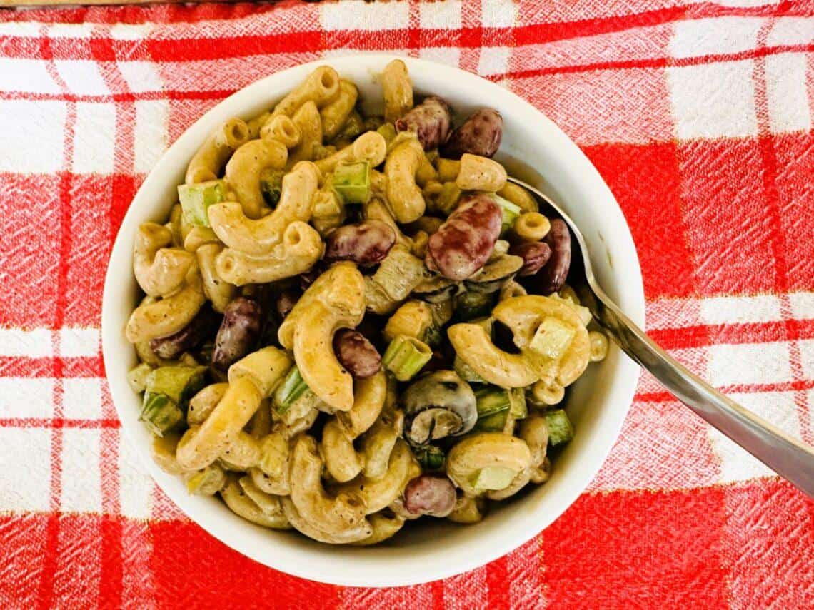 A bowl of macaroni salad with kidney beans, celery, and dressing, perfect for Mexican party food, placed on a red and white checkered cloth with a spoon inside the bowl.