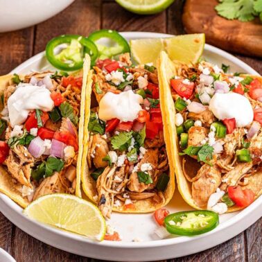 Three chicken tacos topped with diced tomatoes, onions, cilantro, jalapeños, cheese, and sour cream are served on a white plate with lime wedges and surrounded by side dishes—perfect for a flavorful Mexican dinner.