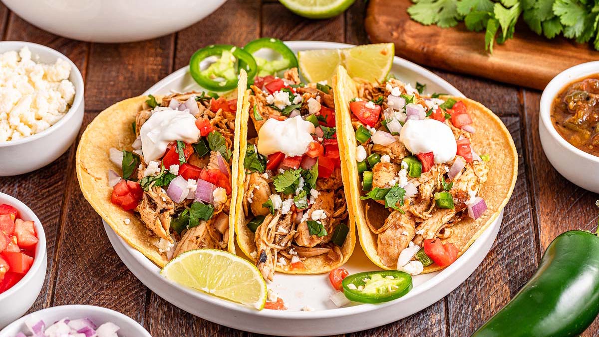 Three chicken tacos topped with diced tomatoes, onions, cilantro, jalapeños, cheese, and sour cream are served on a white plate with lime wedges and surrounded by side dishes—perfect for a flavorful Mexican dinner.