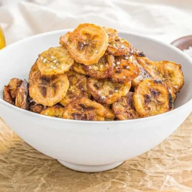 A white bowl filled with golden-brown fried plantain chips, perfect for vegan recipes, with a bottle of oil and a small bowl of salt in the background.