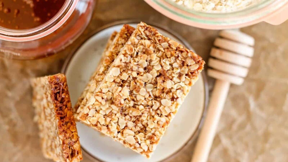 Two oat and honey granola bars sit on a small plate next to a jar of honey and a honey dipper on a parchment surface, making for a healthy dessert option in your collection of oat recipes.