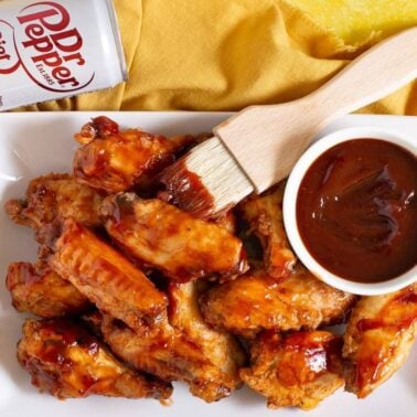 A plate of glazed chicken wings beside a bowl of sauce with a basting brush, and a can of Diet Dr Pepper on a yellow cloth—perfect for easy dinner nights or as a flavorful addition to your favorite slow cooker or crockpot recipes.
