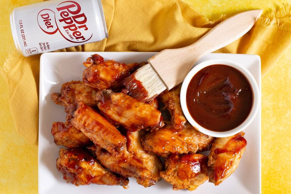 A plate of glazed chicken wings beside a bowl of sauce with a basting brush, and a can of Diet Dr Pepper on a yellow cloth—perfect for easy dinner nights or as a flavorful addition to your favorite slow cooker or crockpot recipes.