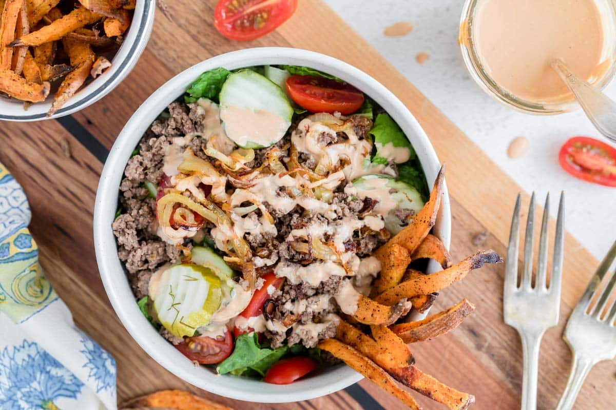 Enjoy one of the best easy bowl meals: a vibrant salad with ground beef, lettuce, pickles, cherry tomatoes, caramelized onions, sweet potato fries, and creamy dressing, served with a side of fries and a glass of sauce.