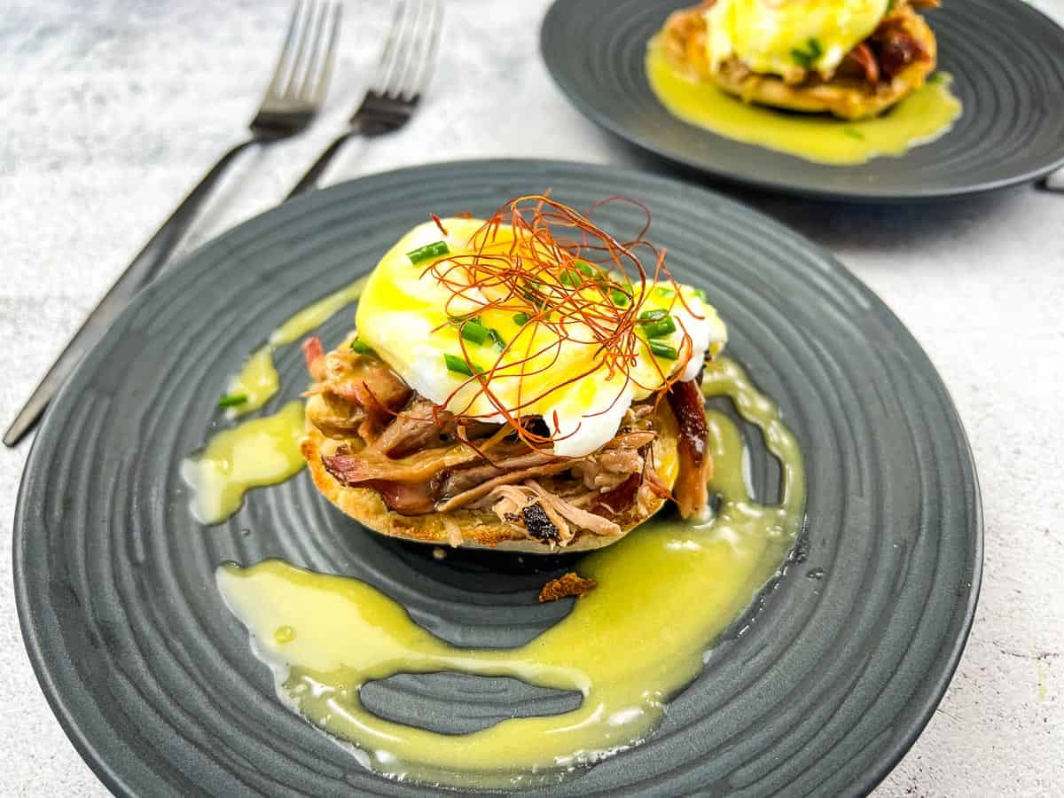 A poached egg on shredded meat and an English muffin, topped with garnish and yellow sauce, makes for one of those quick breakfasts, served on a dark plate with a fork and knife nearby.