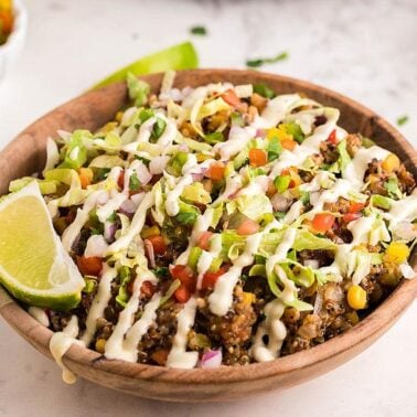 A wooden bowl filled with a colorful quinoa salad topped with diced vegetables, avocado, creamy dressing, and a lime wedge, on a white surface—a perfect example of tasty bowl dishes with fresh lime wedges and herbs.