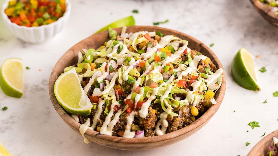 A wooden bowl filled with a colorful quinoa salad topped with diced vegetables, avocado, creamy dressing, and a lime wedge, on a white surface—a perfect example of tasty bowl dishes with fresh lime wedges and herbs.