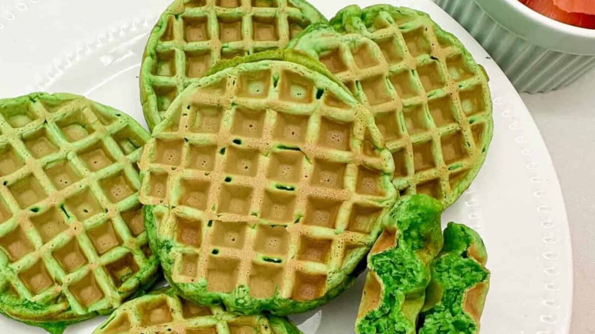 A plate of round, green waffles with a crispy texture, one broken in half to show the inside—perfect for quick breakfasts. A bowl of fruit is partially visible in the background.