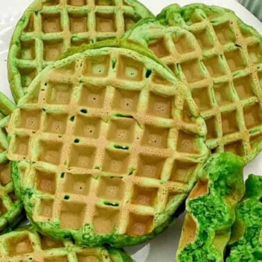 A plate of round, green waffles with a crispy texture, one broken in half to show the insideโperfect for quick breakfasts. A bowl of fruit is partially visible in the background.