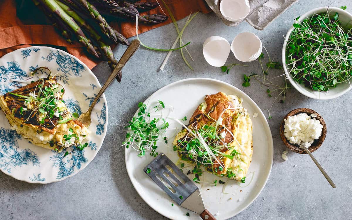 Two plates with slices of omelette topped with microgreens, surrounded by fresh asparagus, eggshells, ricotta cheese, and a bowl of microgreens—perfect inspiration for easy omelet recipes and delicious omelet variations to cook omelets at home.