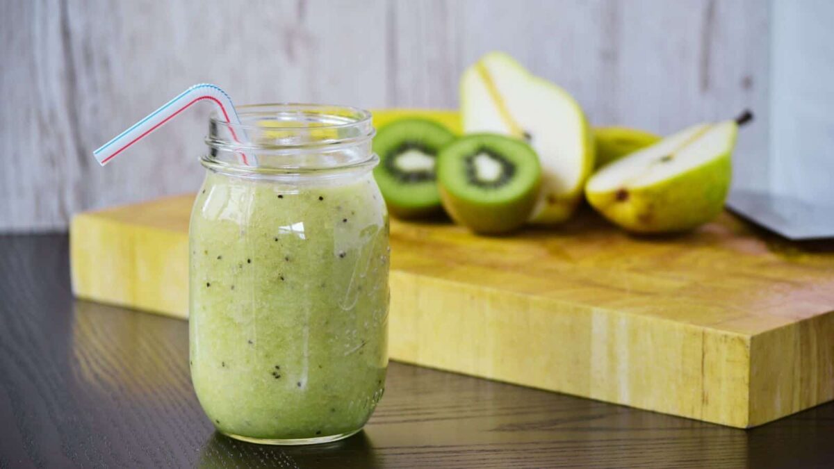 A mason jar filled with a green kiwi and pear smoothie with a striped straw, set on a table; sliced pears and kiwi on a wooden cutting board in the background—perfect for healthy smoothies or as a refreshing meal replacement.