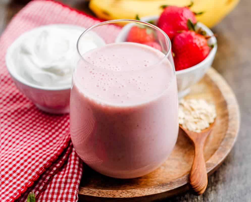 A glass of pink smoothie on a wooden tray, with a bowl of whipped cream, fresh strawberries, oats on a spoon, and a red checkered cloth nearby—perfect for those seeking healthy smoothies or delicious meal replacement smoothies.
