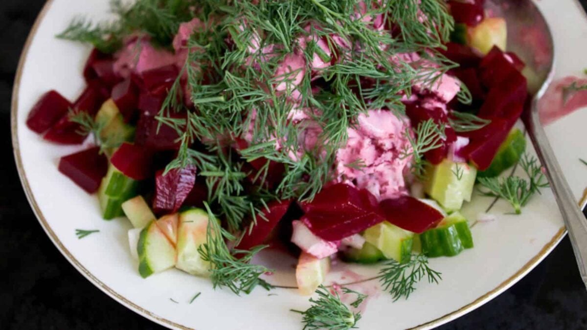 A plate of salad with diced beets, cucumbers, onions, creamy dressing, and fresh dill on top—a classic touch of European cuisine. A spoon rests on the side.