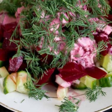 A plate of salad with diced beets, cucumbers, onions, creamy dressing, and fresh dill on top—a classic touch of European cuisine. A spoon rests on the side.