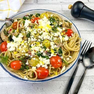 A bowl of spaghetti topped with cherry tomatoes, greens, crumbled cheese, and herbs sits on a white table—an inviting choice for affordable meals—beside a fork, spoon, pepper grinder, and yellow-checked napkin.