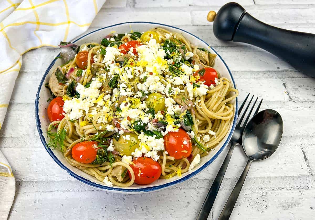 A bowl of spaghetti topped with cherry tomatoes, greens, crumbled cheese, and herbs sits on a white table—an inviting choice for affordable meals—beside a fork, spoon, pepper grinder, and yellow-checked napkin.