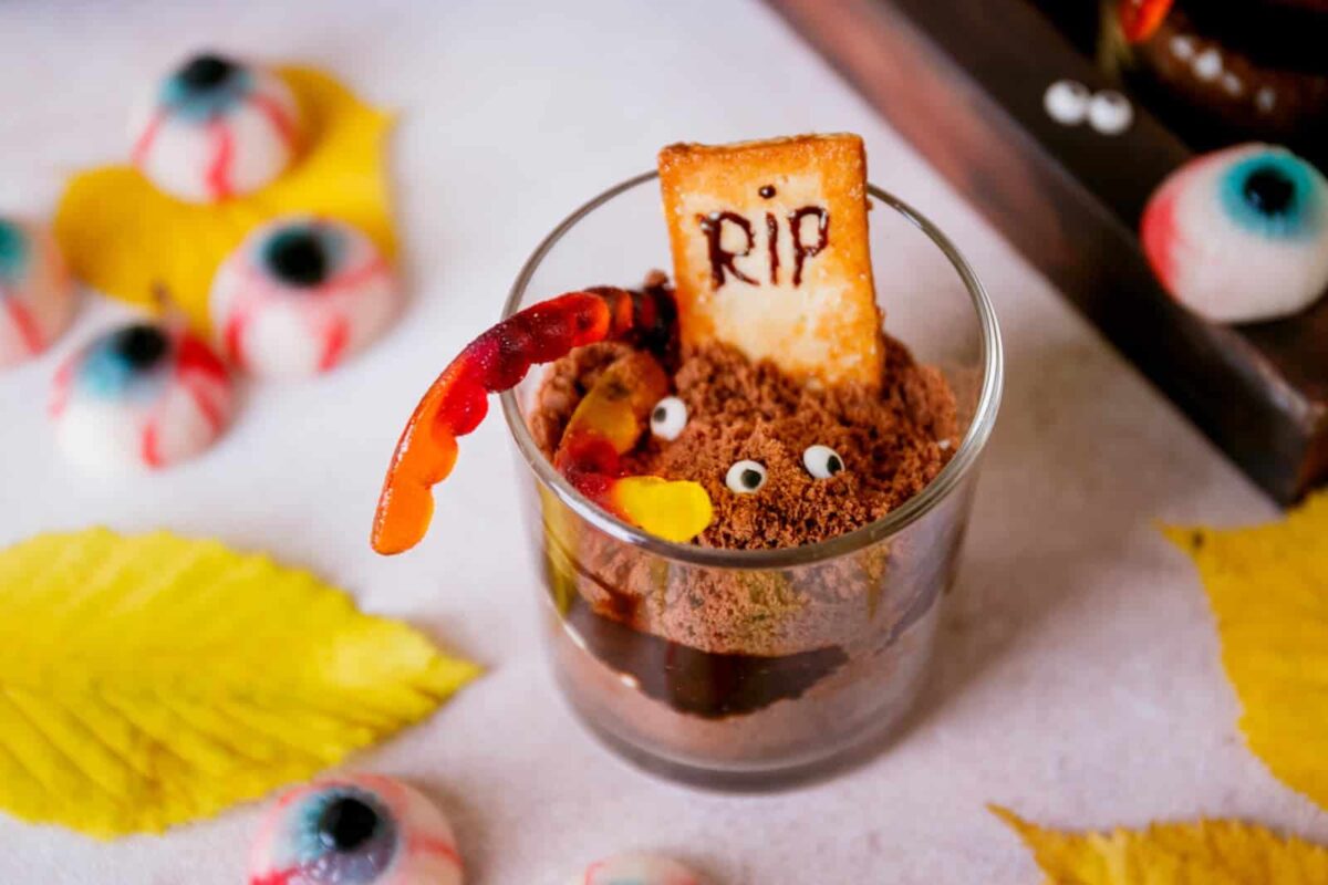 A glass cup with chocolate dessert is decorated with a cookie labeled "RIP," candy eyes, a gummy worm, and candy eyeballs—perfect for spooky recipes and surrounded by yellow leaves.