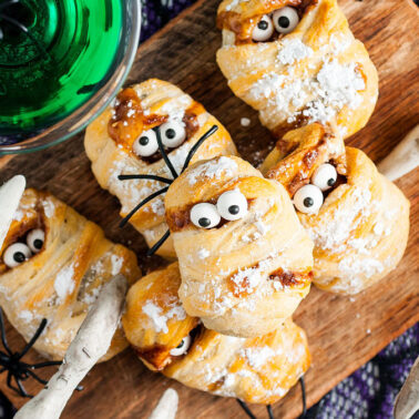 A wooden board with spooky, mummy-shaped pastries topped with candy eyes and powdered sugar, surrounded by decorative white branches and a glass of green liquid—Halloween treats that are fun to make and serve.