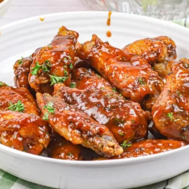 A white bowl filled with flavorful chicken wings coated in sticky barbecue sauce, garnished with chopped herbs, sits on a plaid cloth.