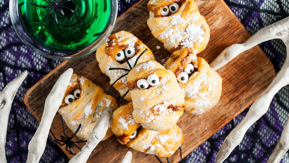 A wooden board with spooky, mummy-shaped pastries topped with candy eyes and powdered sugar, surrounded by decorative white branches and a glass of green liquid—Halloween treats that are fun to make and serve.