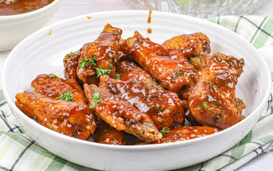 A white bowl filled with flavorful chicken wings coated in sticky barbecue sauce, garnished with chopped herbs, sits on a plaid cloth.