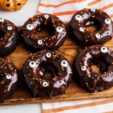 Six chocolate-glazed donuts decorated with candy eyes and orange and black sprinkles are arranged on a wooden board, making these a perfect addition to your collection of spooky recipes, with a striped towel and pumpkin decorations in the background.
