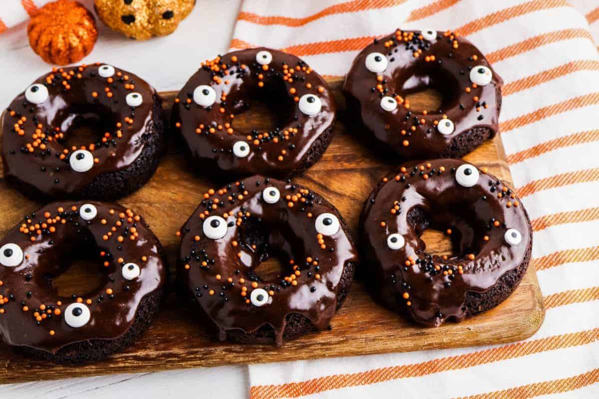 Six chocolate-glazed donuts decorated with candy eyes and orange and black sprinkles are arranged on a wooden board, making these a perfect addition to your collection of spooky recipes, with a striped towel and pumpkin decorations in the background.