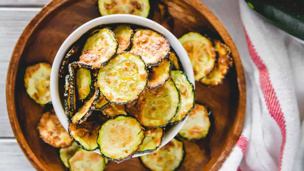 A bowl of baked zucchini chips, perfect for easy appetizers or air fryer recipes, sits inside a larger wooden bowl filled with more chips, next to a white towel with red stripes.