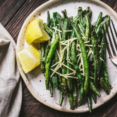 A plate of roasted green beans topped with shredded cheese, served with two lemon wedges and a fork on a wooden table—perfect for easy appetizers or as a delicious addition to your air fryer recipes collection.