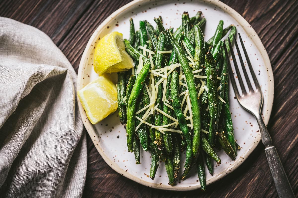 A plate of roasted green beans topped with shredded cheese, served with two lemon wedges and a fork on a wooden table—perfect for easy appetizers or as a delicious addition to your air fryer recipes collection.