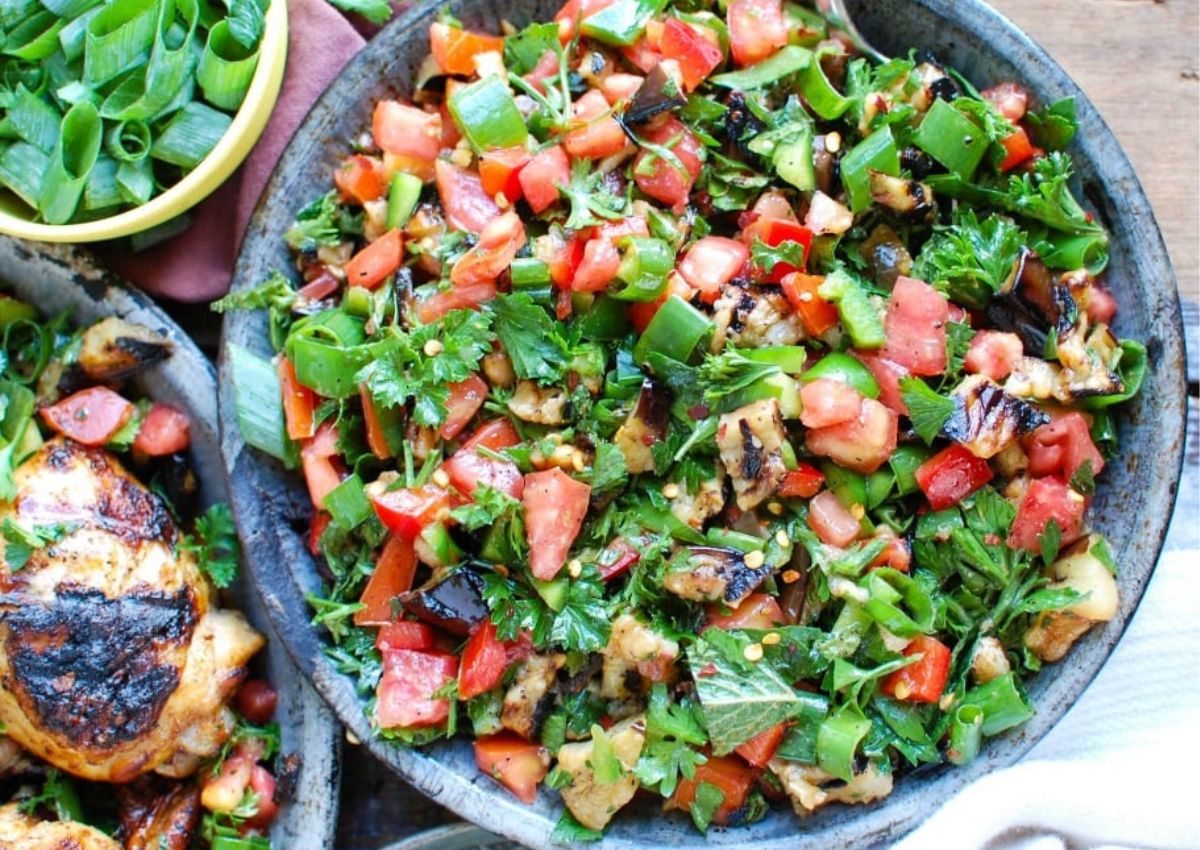 A bowl of chopped grilled vegetables, including eggplant, tomatoes, green onions, cilantro, and herbs is shown on a plate next to a bowl of sliced green onions and grilled meat—perfect inspiration for flavorful eggplant dishes or creative dinner recipes.