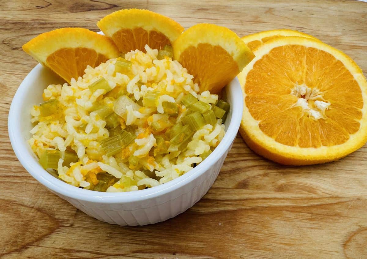 A white bowl of rice mixed with chopped vegetables, garnished with orange slices, sits on a wooden surface next to additional sliced oranges—perfect for those seeking creative rice side dishes.