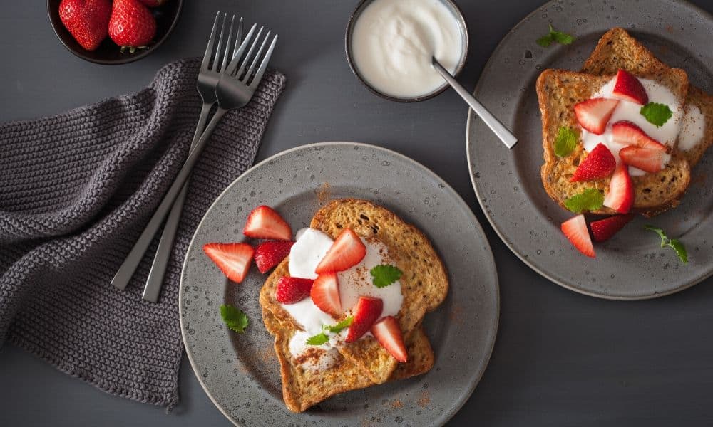 Two plates of sweet French toast topped with sliced strawberries, yogurt, and herbs sit beside a bowl of strawberries, a bowl of yogurt, two forks, and a gray napkin on a dark surface.
