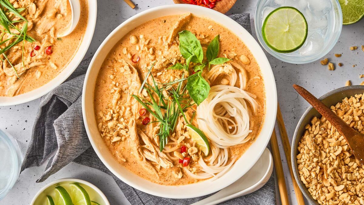 A bowl of Thai-inspired noodle soup with creamy broth, garnished with herbs, lime, peanuts, and chili—perfect comfort food—surrounded by extra lime, peanuts, and a glass of water. One of the coziest soups to enjoy anytime.
