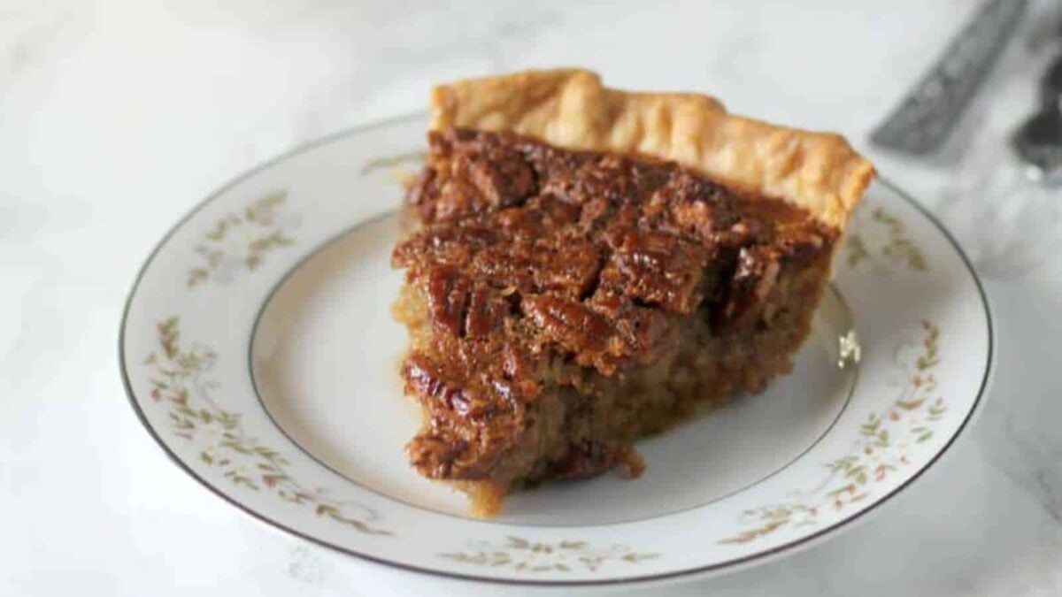 A slice of pecan pie sits on a decorative white plate with a floral pattern, placed on a light-colored surface—an inviting treat among classic Thanksgiving desserts.